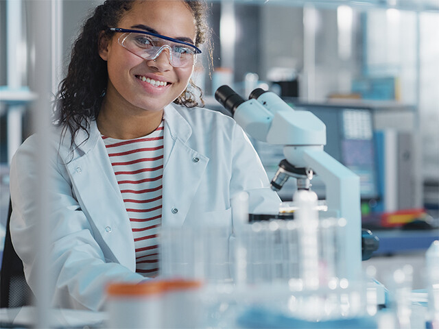 young woman in white lab coat smiling behind a microscope