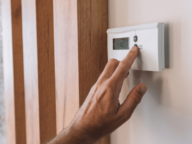 Detail ruky nastavující digitální pokojový termostat na stěně. Ovládání slouží k regulaci teploty v interiéru a optimalizaci výkonu systému podlahového vytápění.