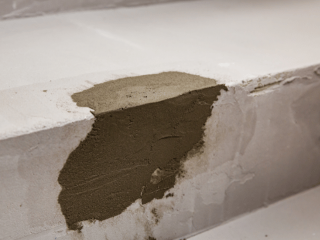 Detail opravy poškozené hrany schodu pomocí cementové opravné malty. Materiál je nanesen na poškozené místo pro obnovení tvaru a pevnosti betonového povrchu.