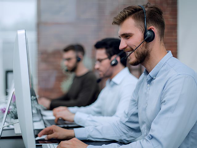 Tre personer med headset arbejder ved computere på et kontor. Personen i forgrunden sidder ved en skrivebordsskærm, mens to kolleger arbejder ved arbejdsstationer i baggrunden.