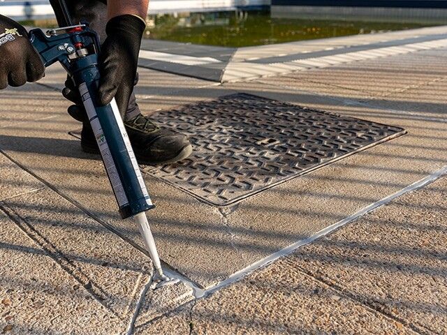 Bostik chemical-resistant sealant being applied with a cartridge gun into expansion joints between concrete paving slabs in an outdoor area.