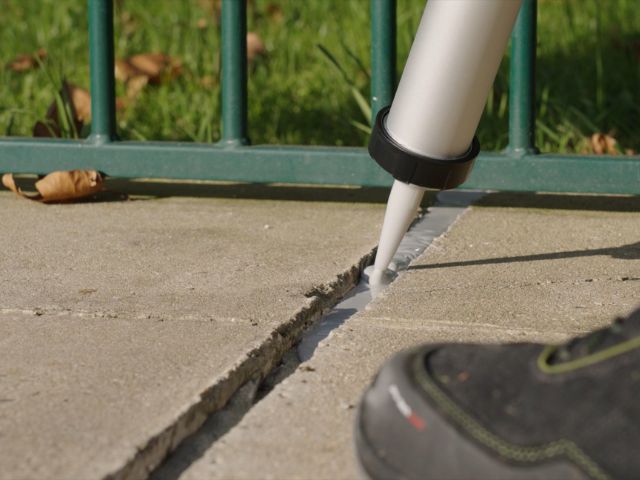 Bostik chemical-resistant sealant being applied with a cartridge gun into an expansion joint between concrete paving slabs in an outdoor area.