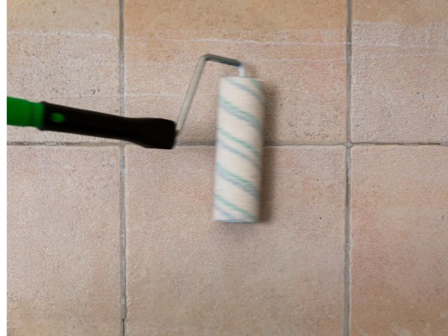 Paint roller applying Bostik primer evenly across a tiled floor surface, preparing the substrate before tiling.