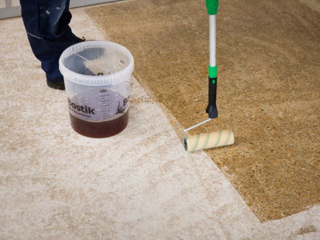 Bostik primer being applied with a roller next to a bucket on a prepared subfloor, showing transition between untreated and primed surfaces.