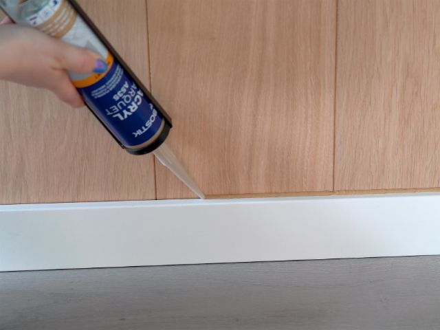 Bostik acrylic sealant being applied along the junction between a white skirting board and wood-effect wall panelling to create a neat, flexible seal.