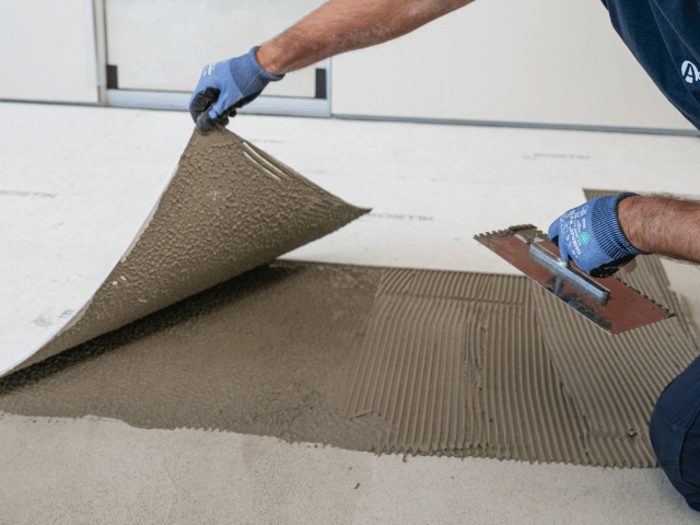 Bostik floor adhesive being spread with a notched trowel while installing floor panels on a prepared concrete subfloor.