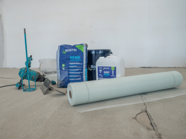 Bostik Reno U550 system materials and tools arranged on a concrete floor, including primer, repair compound, mixing bucket, tools, and reinforcement mesh roll.