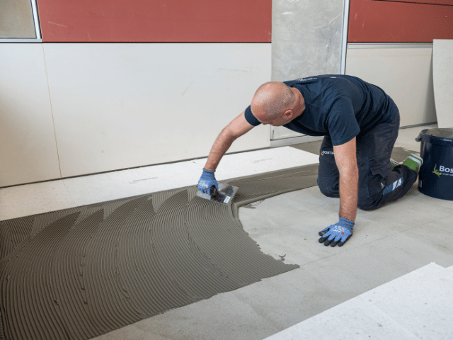 Bostik floor adhesive being evenly spread with a notched trowel across a concrete subfloor to prepare for installation of flooring panels.