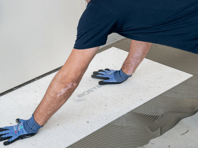 Bostik floor panel being positioned onto combed adhesive using gloved hands to ensure proper placement during installation.