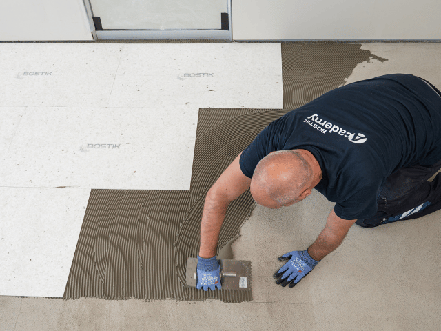 Bostik floor adhesive being spread with a notched trowel while installing floor panels in a staggered pattern on a concrete subfloor.