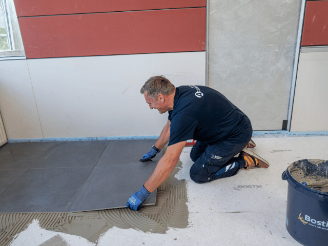 Large floor tiles being laid into Bostik adhesive on a prepared subfloor, ensuring even placement during flooring installation.