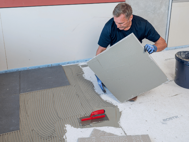Large floor tile being positioned into Bostik adhesive on a prepared subfloor, with a notched trowel and adhesive layer visible during installation.
