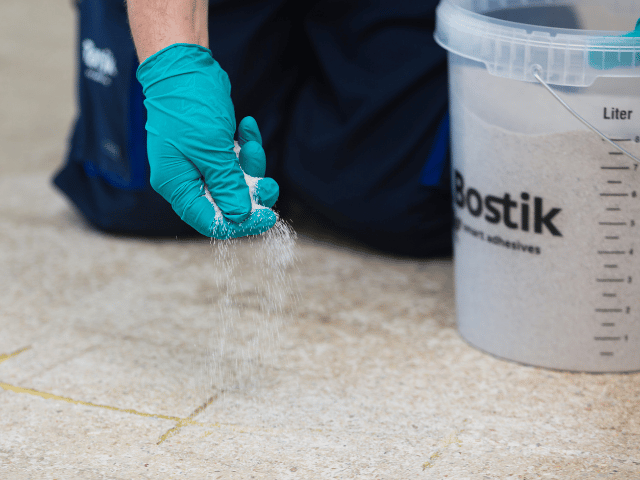Bostik repair sand being sprinkled by hand onto treated cracks on a concrete floor to enhance bonding and surface stability.