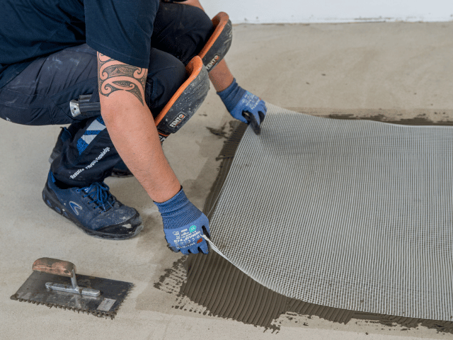 Bostik reinforcement mesh being positioned into adhesive on a concrete subfloor using a notched trowel for even coverage.