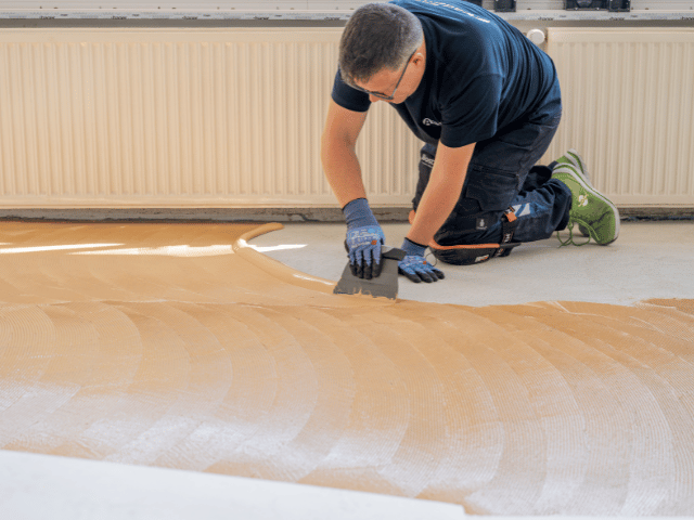 Bostik floor adhesive being spread evenly with a trowel across a concrete subfloor during renovation preparation.