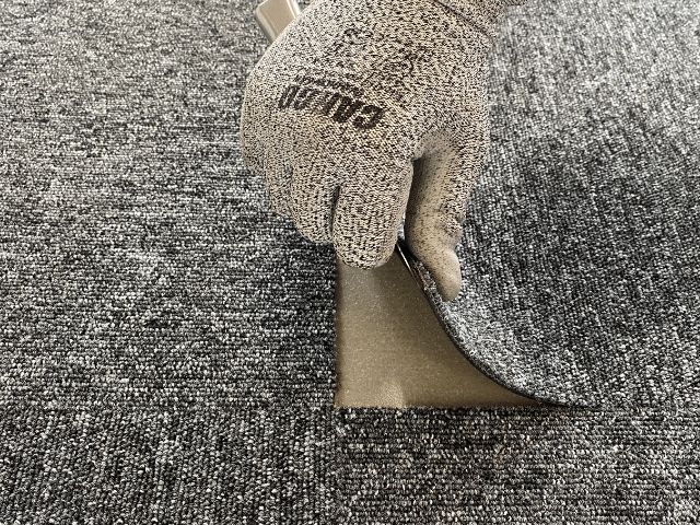 Gloved hand lifting a carpet tile to reveal adhesive underneath, demonstrating flooring installation using Bostik solutions.