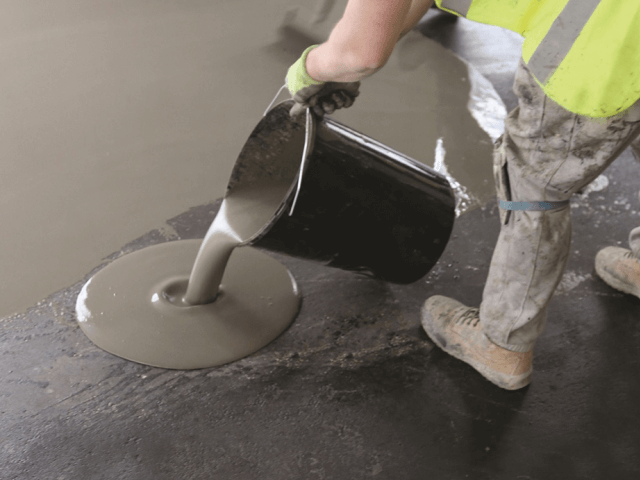 Worker pouring self‑levelling compound from a bucket onto a concrete floor to create a smooth surface.