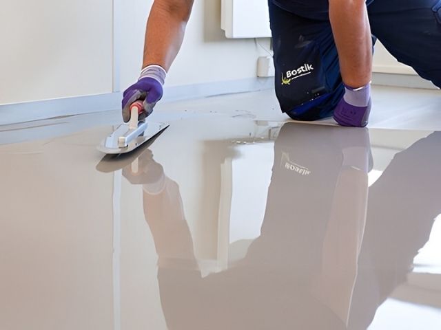 Worker smoothing a self‑levelling floor compound with a trowel to create an even, glossy surface indoors.