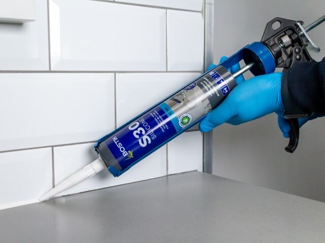 A gloved hand holds a caulking gun with silicone sealant, applying it to the joint between a countertop and white tile backsplash