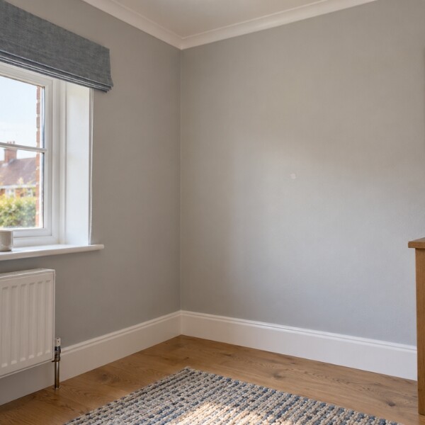 Light grey painted interior wall in the corner of a room with a small white repair patch visible on the surface. The room features white skirting boards, a wooden floor, a patterned rug, a window with a grey fabric blind, and a white radiator beneath the window.