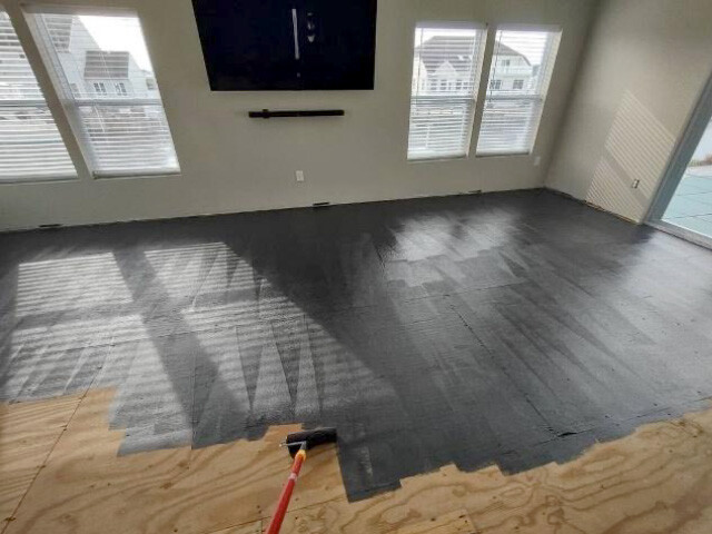 An empty living room with Bostik Roll-Cote black roll-on underlayment over plywood subfloor.