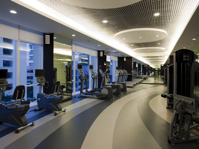 A room with gym equipment, such as treadmills, ellipticals, and weight stations, sitting on multi colored rubber floors.