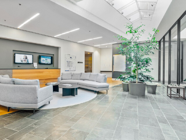 A waiting room with tile flooring, two gray sofas, a potted tree and two televisions.