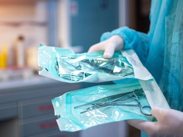 A medical professional in surgical PPE holds pre-packaged sterilized medical equipment in his hands. 